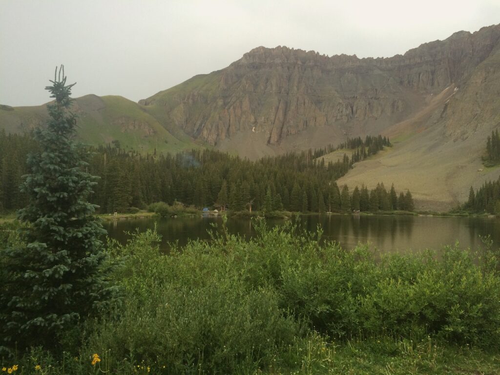 Alpine lake surrounded by pine trees with mountain views and distant campers in background. Alta Lakes, Telluride, Coloardo.