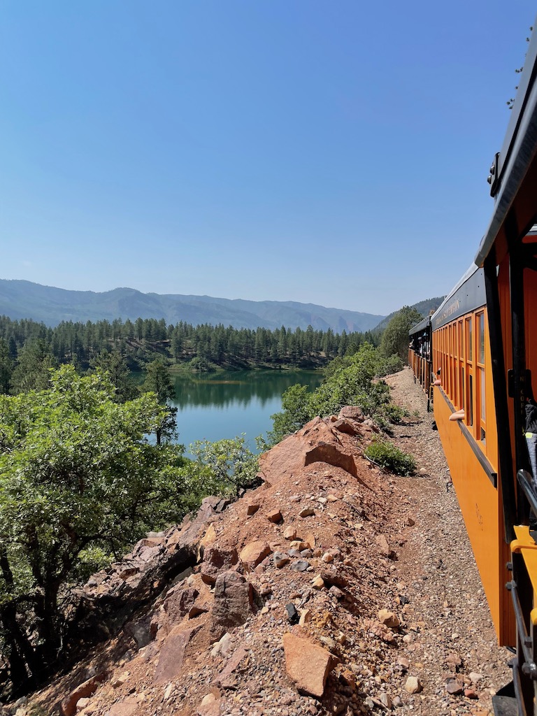 Train winding through mountains alongside alpine lake with mountain views in background on cloudless summer day. Durango Silverton Train, Rocky Mountains, Durango, Colorado.