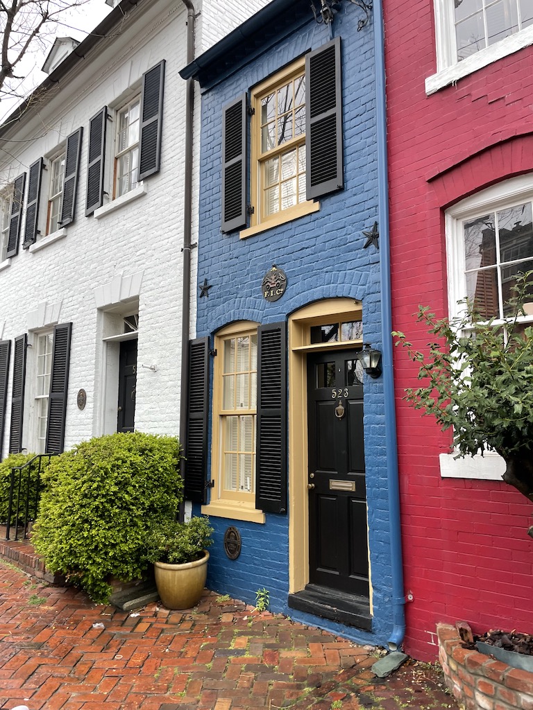 White, blue, and red painted brick townhomes with extremely narrow townhome in center. Historic homes and brick sidewalks. Old Town Alexandria, Virginia, Washington DC.