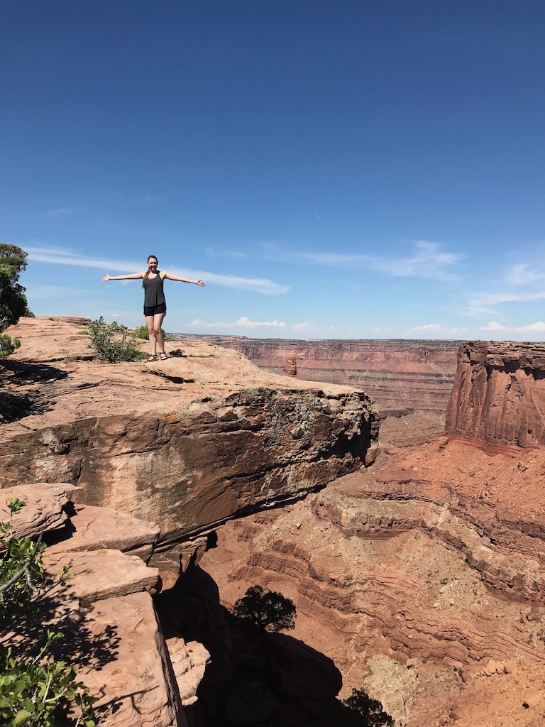 Woman standing on desert rock cliff edge with red rock canyons in background on sunny summer day. Dead Horse Point State Park, Moab, Utah.