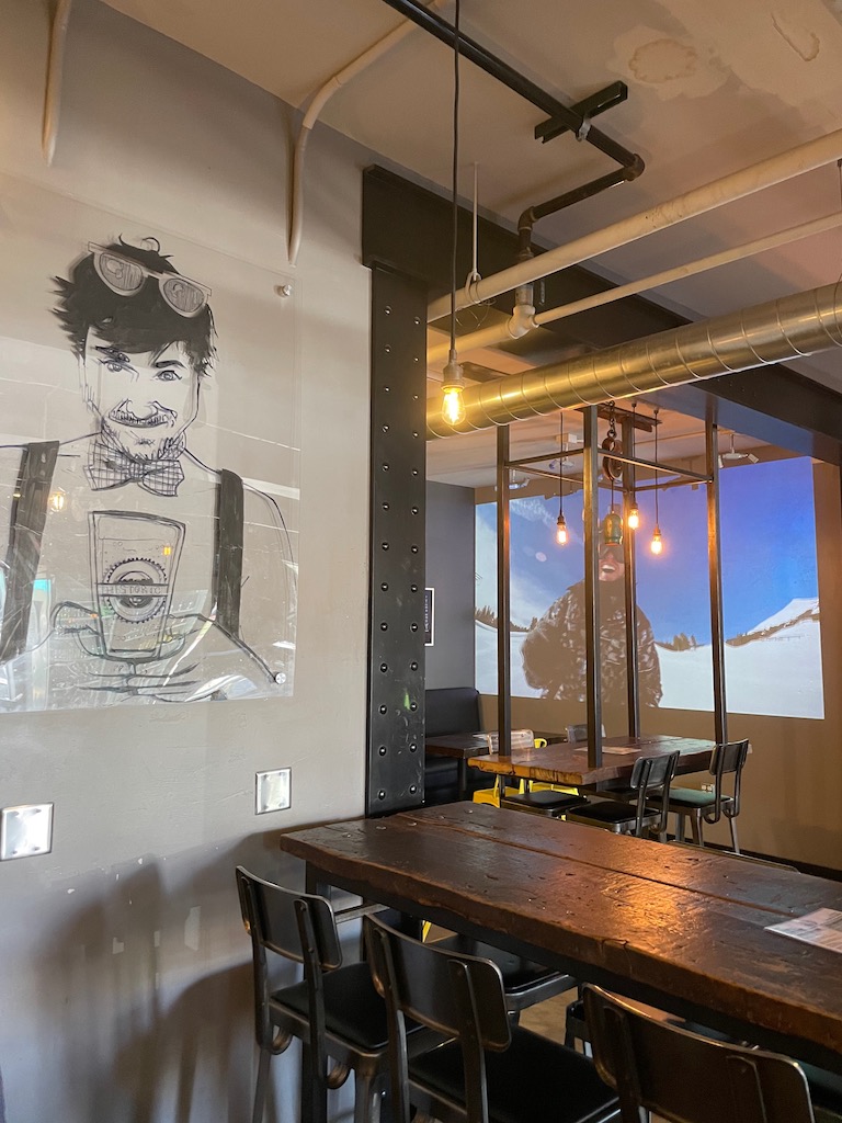 Modern restaurant with wood tables, metal chairs, acrylic artwork with drawing of man holding beer, in background is large projection screen showing snowboarding videos. Historic Brewing Barrel and Bottle House, downtown Flagstaff, Arizona.