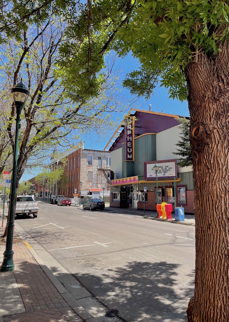 Downtown street lined with low brick buildings, brick-lined sidewalks, and mature trees, with colorful building in center with large sign reading "ORPHEUM". Orpheum Theater, Historic Downtown Flagstaff, Arizona.