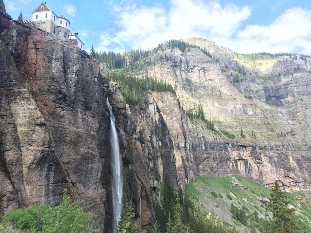 300 foot free falling waterfall with small, historic electric house at top, waterfall is surrounded by sheer rocky cliffs and greenery at base on sunny summer day. Bridal Veil Falls, San Juan Mountains, Telluride, Colorado.