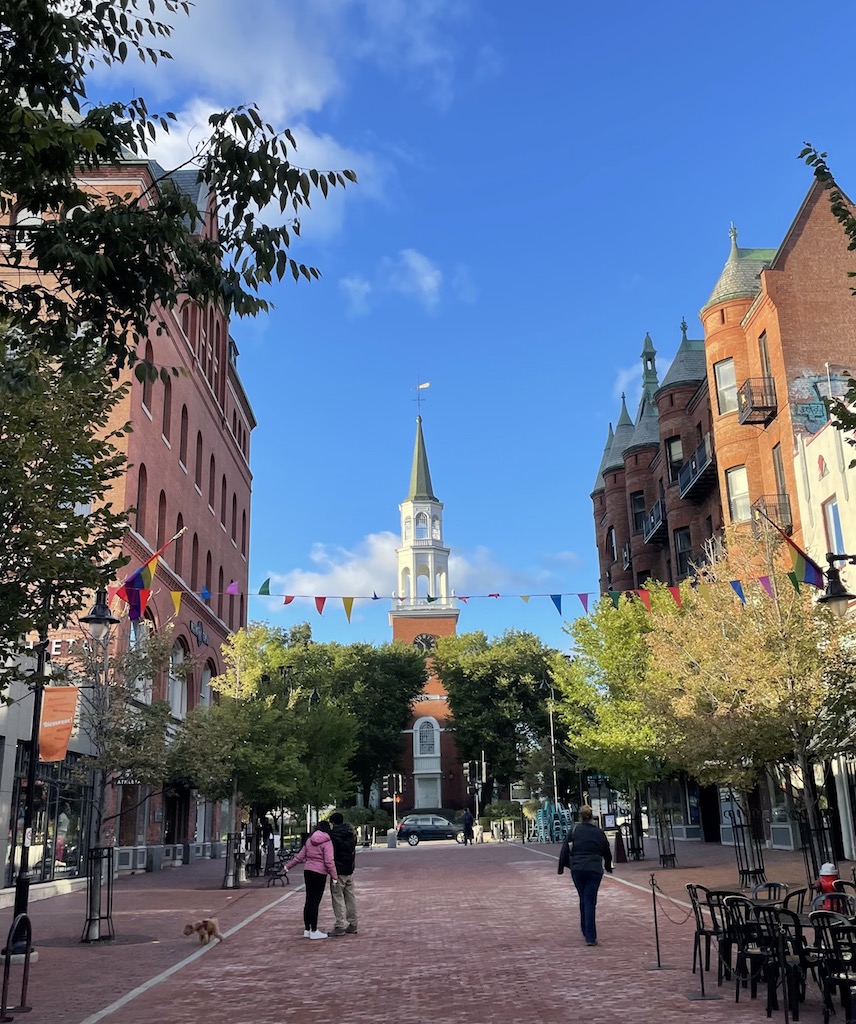Brick pedestrian sidewalk lined with brick buildings and sidewalks tables leading to statuesque church with tall steeple at end of walk way. Church Street, Burlington, Vermont, New England.