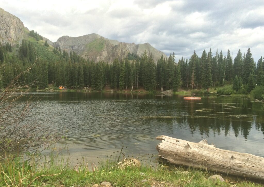 Man kayaking on calm alpine lake surrounded by pine trees and mountain views on cloudy summer evening. Alta Lakes, San Juan Mountains, Telluride, Colorado.