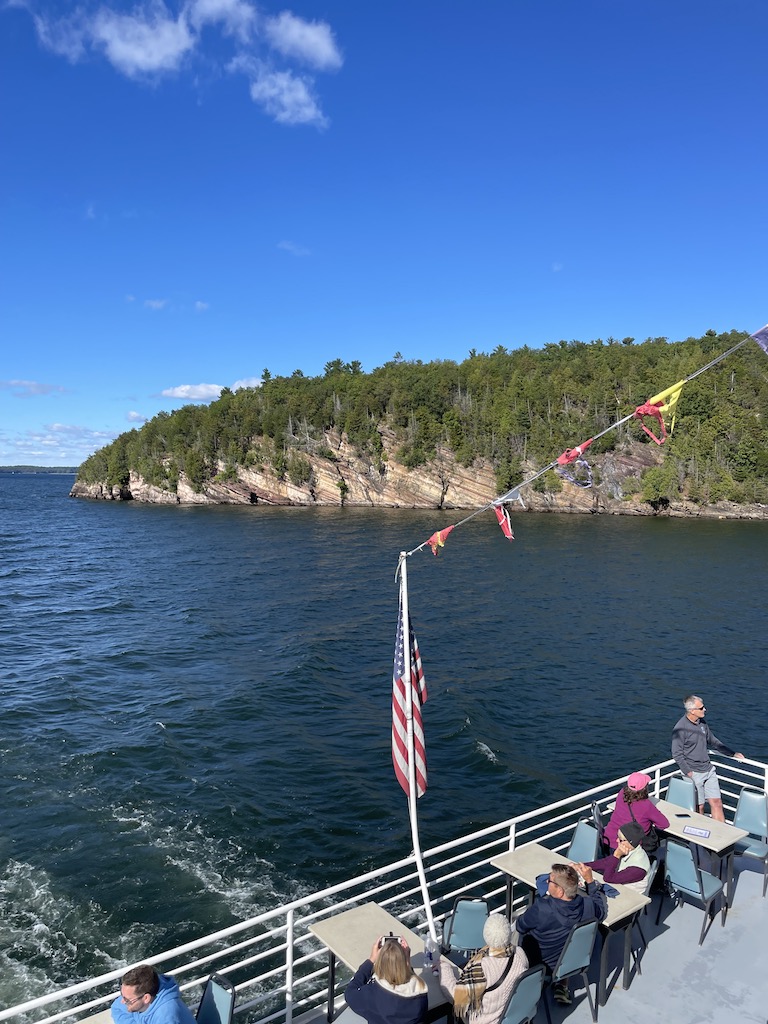 Large boat with people sitting at chairs and tables overlooking back deck of boat, view is of large lake with tree-covered rocky cliffs in background on a sunny summer day. Boat tour, Lake Champlain, Burlington, Vermont, New England.