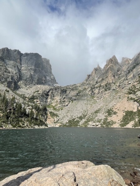 Small mountain lake with dark blue water and small choppy waves surrounded by towering, rocky peaks and occasional pine trees on cloudy and windy summer day. Emerald Lake hike, Rocky Mountain National Park, Estes Park, Colorado.