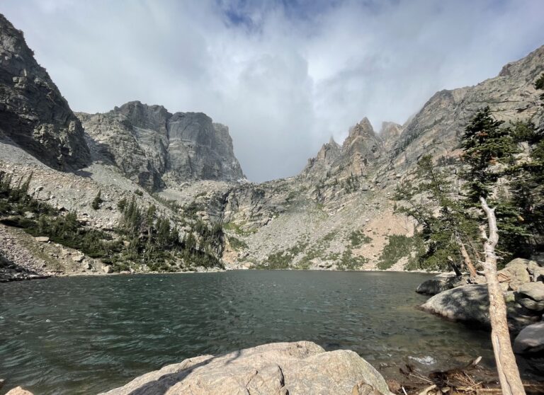 Small mountain lake with dark blue water and small choppy waves surrounded by towering, rocky peaks and occasional pine trees on cloudy and windy summer day. Emerald Lake hike, Rocky Mountain National Park, Estes Park, Colorado.