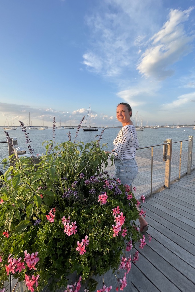 Woman smiling at camera on dock overlooking sailboats and yachts on Sag Harbor during sunny summer evening, with pink and purple flower pots in foreground. Sag Harbor Yacht Yard, Sag Harbor, New York, The Hamptons. How I quit my job and traveled for a year.