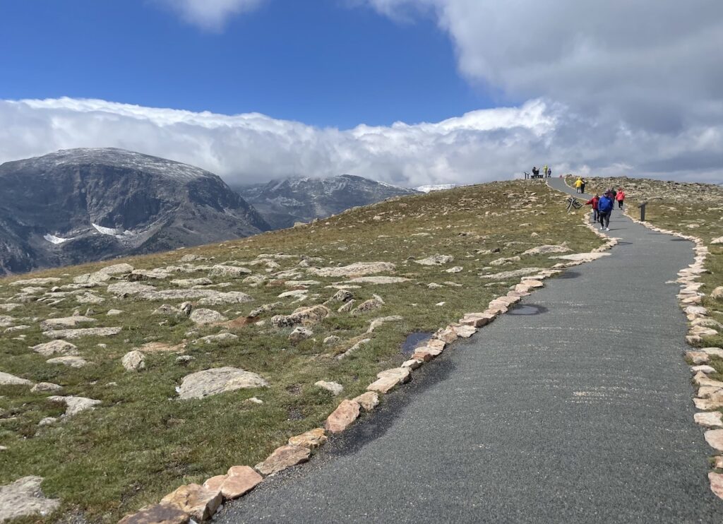 People walking up paved path on mountain tundra hill with huge, dramatic mountain-scapes in background on sunny summer day. Trail Ridge Road, Rocky Mountain National Park, Estes Park, Grand Lake, Colorado.