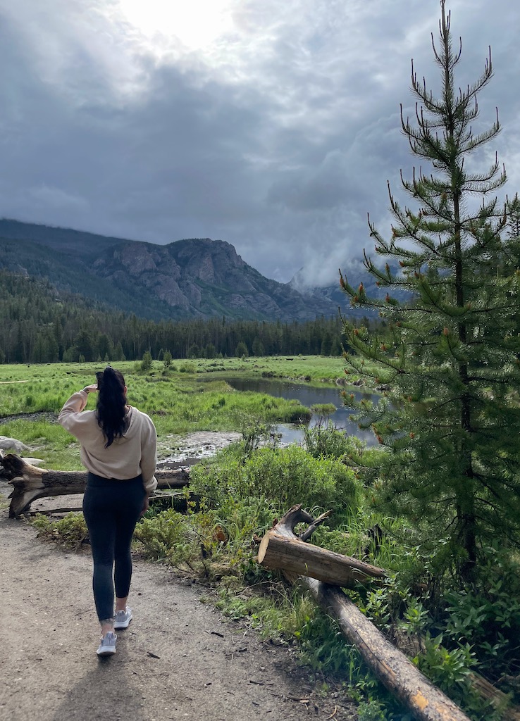 Woman on dirt hiking trail overlooking green meadow with calm river and mountain views in background on cloudy summer morning in Rocky Mountain National Park. East Inlet trail, Rocky Mountain National Park west side, near Grand Lake, Colorado.