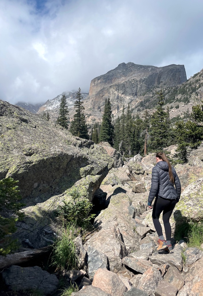 Woman hiking on rocky boulder path with pine trees and rocky mountains in background on cloudy summer day. Lake Haiyaha hiking trail, Rocky Mountain National Park, near Estes Park, Colorado.