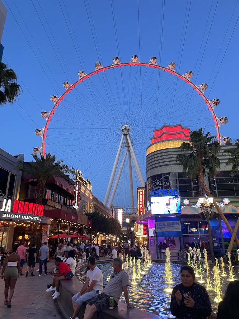 Huge observation wheel / ferris wheel lit up in red at night surrounded by colorfully lit buildings, fountains, and crowds of people. The High Roller ferris wheel at the Linq, Las Vegas strip at night, Nevada.