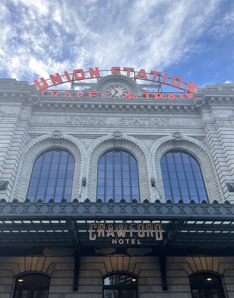 Historic building with large arched windows, ornate framing, and huge neon sign reading "Union Station Travel by Train" on sunny day. Historic Denver Union Station, Denver, Colorado.