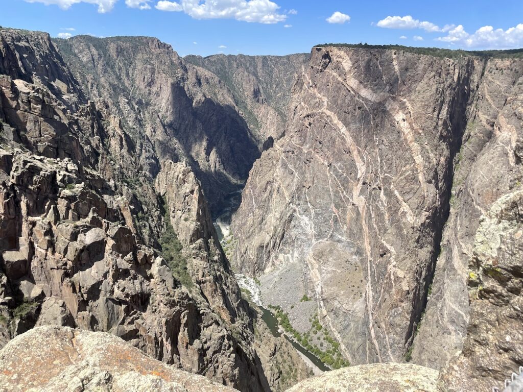 Huge rock cliff, taller than almost any sky scraper in the world, cliff face is almost vertical, brown, with white stripes, below the canyon walls is a narrow river. Painted Wall, Black Canyon of the Gunnison National Park, South Rim, near Montrose, Colorado.