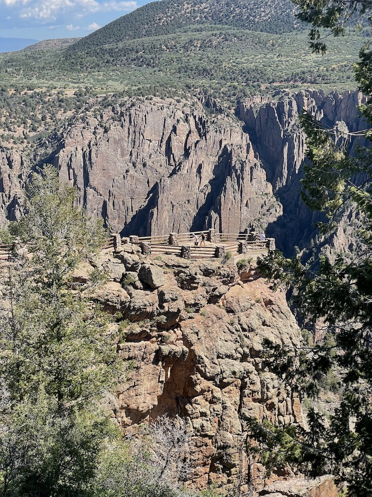 Small overlook jutting out over rocky canyon wall, canyon cliffs are steep, vertical brown jagged rocks, atop the cliff are semi-arid pine forests. Gunnison Point Overlook, Black Canyon of the Gunnison National Park, South Rim, near Montrose, Colorado.