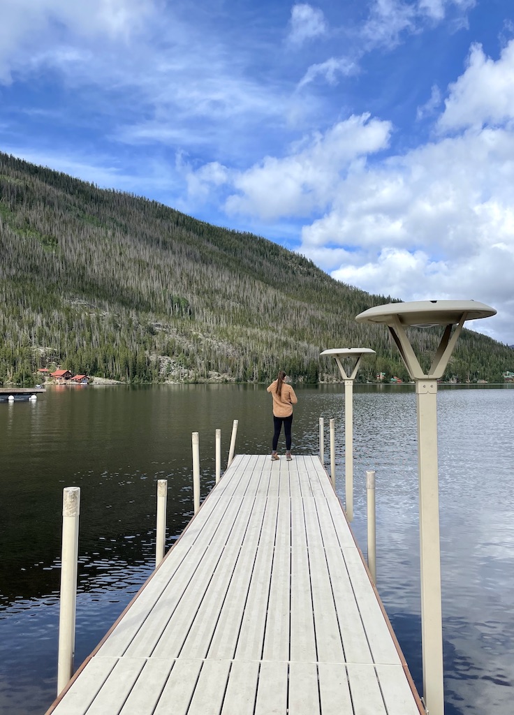 Woman standing on long dock over dark waters of alpine mountain lake with tall mountain peak in background on sunny summer day. Public dock/ boat launch, Grand Lake, Colorado.