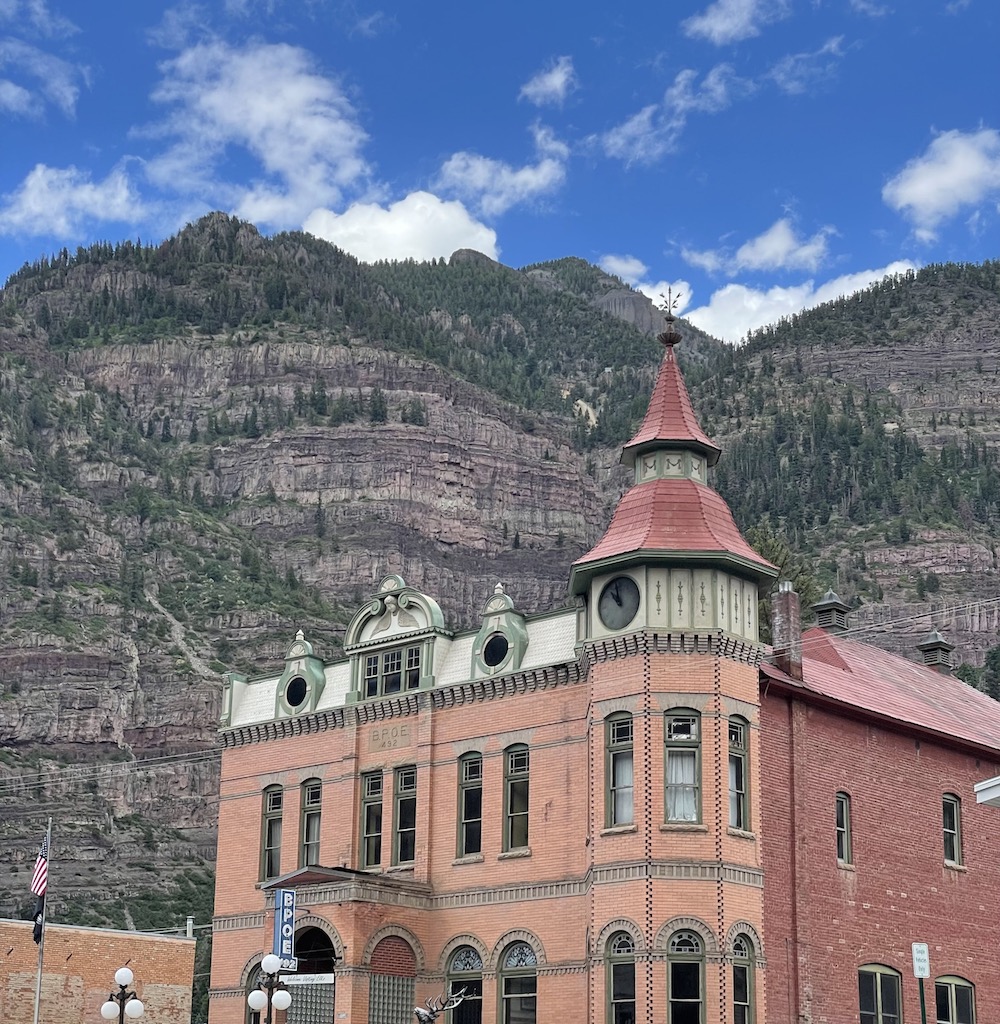 Historic brick building with ornate details, domed tower, and arched windows backed by dramatic mountain views on a sunny summer day. Main Street, Ouray, Colorado.