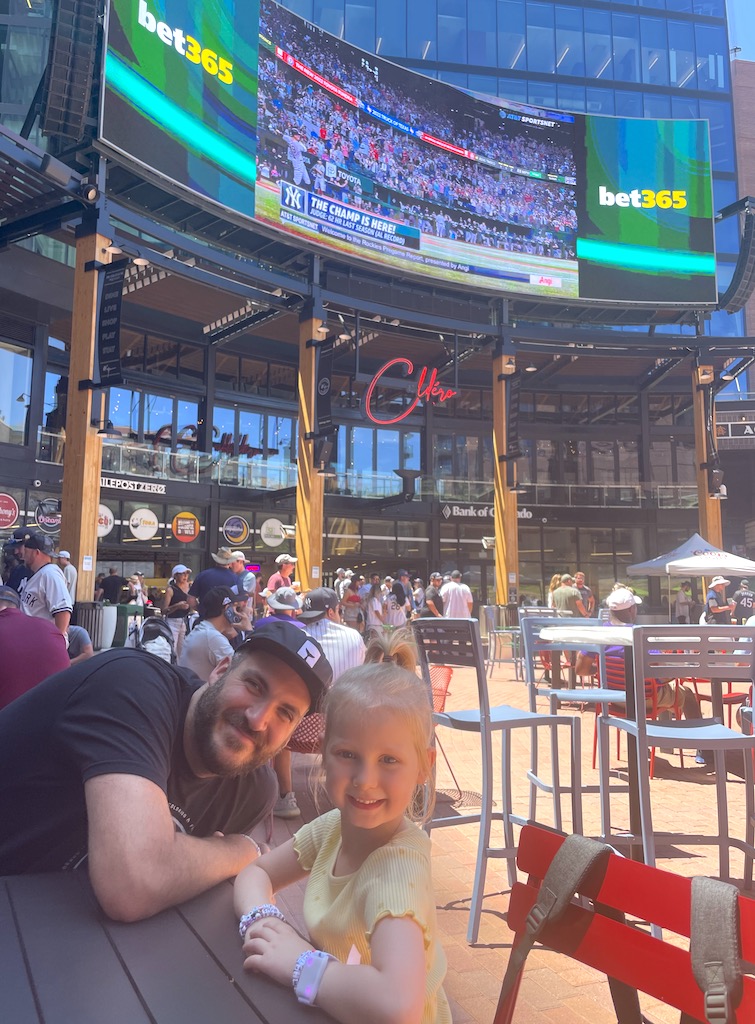 Man and young girl at outdoor bistro table smiling at camera. Bistro table is in large courtyard full of people with huge outdoor tv mounted to high rise building showing baseball game in the back. Sunny summer day in McGregor Square, Denver, Colorado.