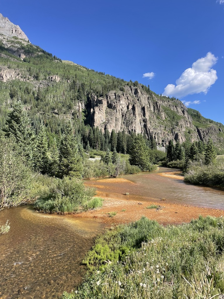 Shallow, wide creek with red sand creek bed surrounded by lush greenery, pine forest, and jagged mountains on sunny summer day. Crystal Lake stop on Million Dollar Highway, Red Mountain Pass, between Silverton and Ouray, Colorado.