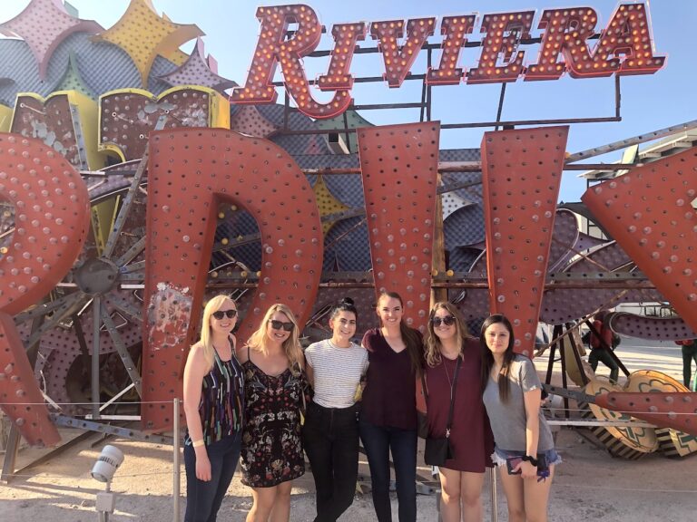 Group of women smiling at camera in front of huge, colorful, old neon signs in Neon Boneyard on sunny day. Neon Boneyard, Neon Museum Las Vegas, Downtown Las Vegas, Las Vegas, Nevada.