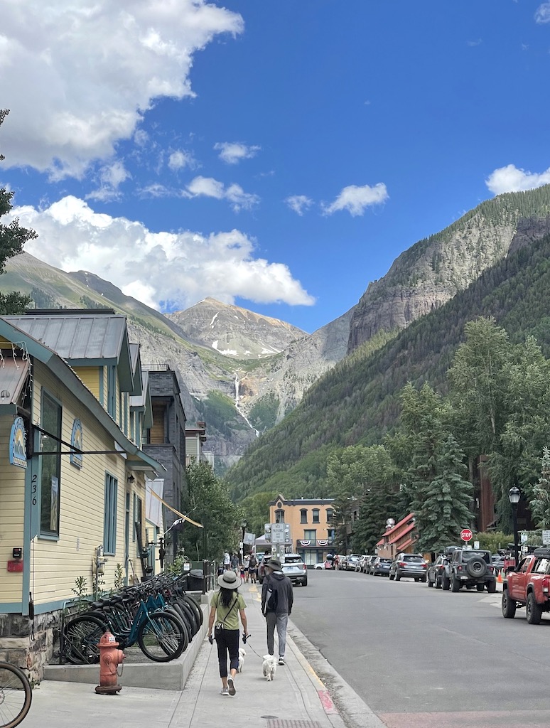 People walking down street lined with historic homes, bikes, and pine trees, in background are stately mountain peaks and distant waterfall. Town of Telluride, Colorado.
