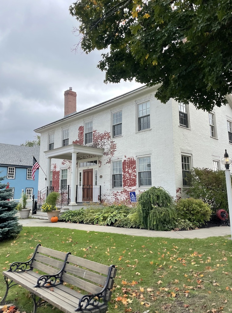 Wooden bench in front of white washed, red brick building with neat lawn decorated with fall decor, flowers, and pumpkins. Historic home in Historic Downtown Stowe, Vermont during fall.