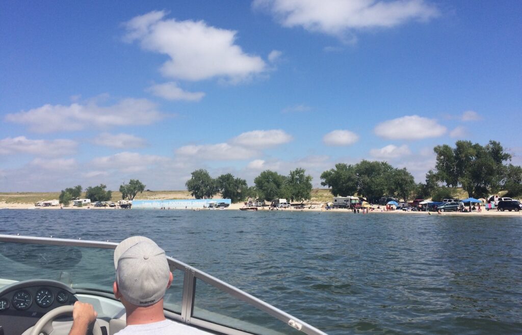 Man driving boat on lake looking out at shoreline covered with rvs and tents, beach campers on sunny summer day. Lake McConaughy, Nebraska.