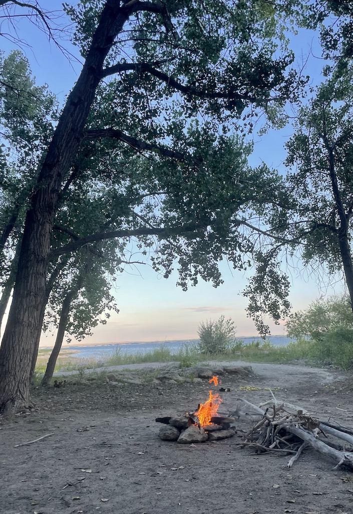Campfire at dirt campsite surrounded by trees with large lake in distance during sunset. Lake McConaughy, Nebraska.