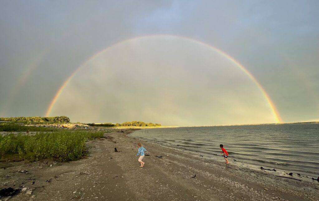 Two kids playing on sandy shoreline of lake during sunset under a huge double rainbow surrounded by storm clouds with sun peaking out in distance. Lake McConaughy, Nebraska.