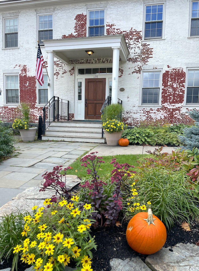 White washed, red brick building with neat lawn decorated with fall decor, flowers, and pumpkins. Historic home in Historic Downtown Stowe, Vermont during fall.