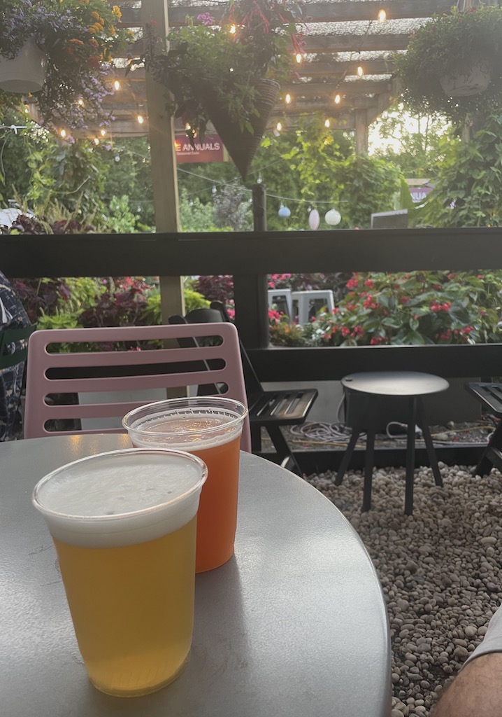 Two craft beers in plastic cups on outdoor bistro table with flowers and trellis with string lights. Kidd Squid Brewing Company, Sag Harbor, New York, The Hamptons.