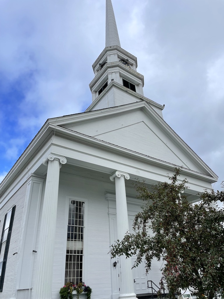 White steepled church with columns, tree in front of church on sunny fall day. Stowe Community Church, Historic Downtown Stowe, Vermont during fall.