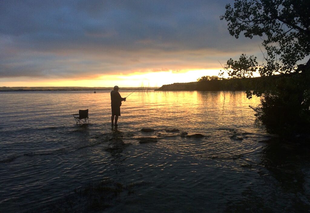 Silhouette of man on shallow shoreline with fishing pole next to camping chair during colorful sunset. Lake McConaughy, Nebraska.