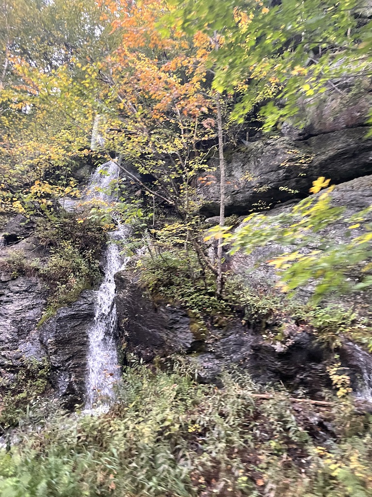 Rocky outcrop with small waterfall surrounded by orange yellow and green fall foliage. Smugglers' Notch Scenic Highway, Mount Mansfield, Stowe, Vermont.