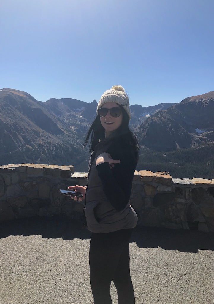 Woman smiling at camera with dramatic mountain views in background on cloudless fall day, woman is wearing beanie hat with sunglasses and long sleeve with puffy vest. Viewpoint on Trail Ridge Road in Rocky Mountain National Park during fall, near Estes Park and Grand Lake, Colorado.