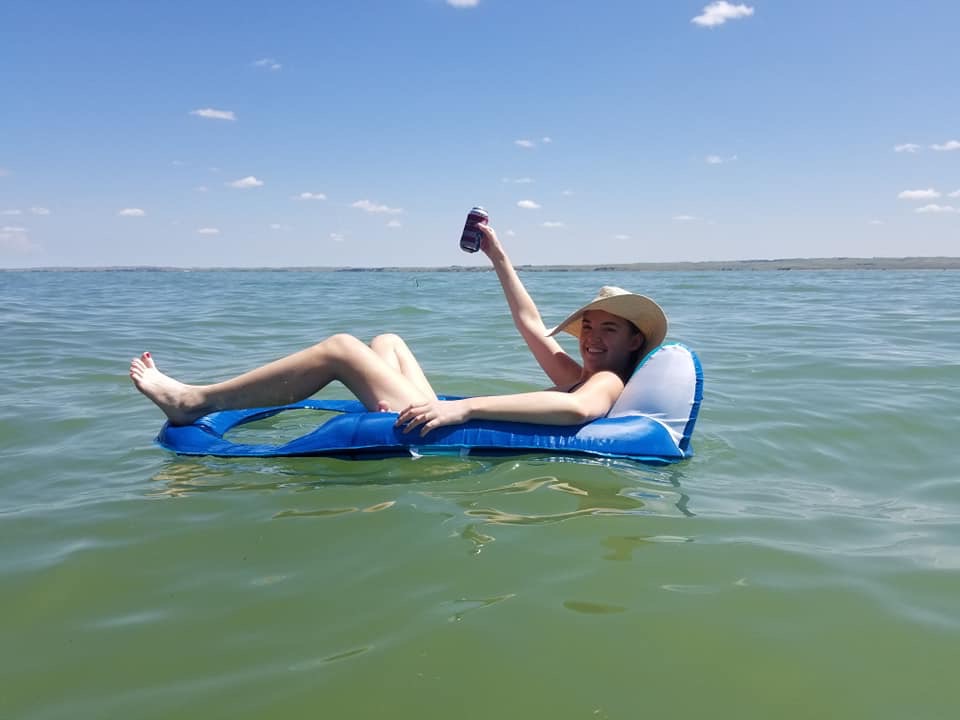 Woman laying on pool float in lake, smiling and raising up drink in her hand during sunny summer day. Lake McConaughy, Nebraska.