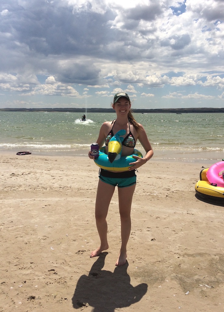 Woman smiling at camera on sandy beach while wearing pool floaty, pool floaties and jetskis in lake in background during sunny summer day. Lake McConaughy, Nebraska.