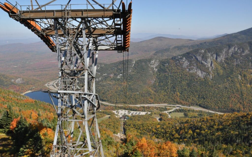 Metal structure of historic aerial tramway, with dramatic mountain and lake views with fall colors of red, orange, green, and yellow on sunny, clear fall day. Cannon Mountain Aerial Tramway, White Mountains, New Hampshire.