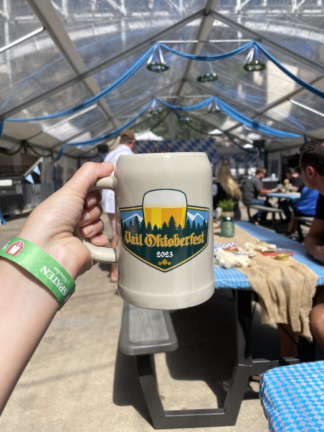 Ceramic beer stein with Vail Oktoberfest logo, with German beer tent and live band in background. Vail Oktoberfest, Vail, Colorado.