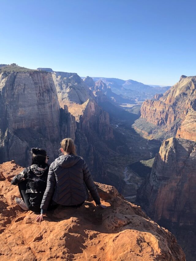 Two women sitting on rocky cliff edge overlooking dramatic views of Zion Canyon, filled with red rocks and lush greenery on cloudless fall day. Observation Point Trail, Zion National Park, Utah.