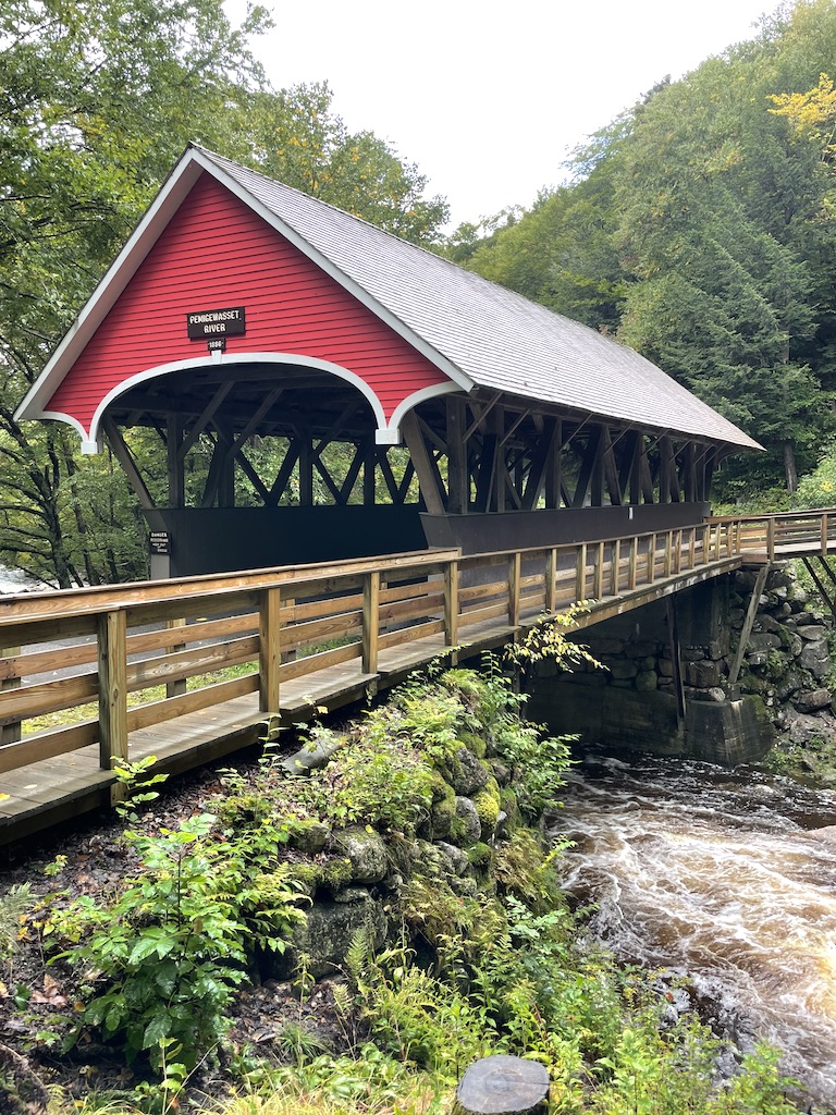 Red wooden covered bridge over river, surrounded by lush green trees in the mountains on cloudy day. Flume Bridge, Flume Gorge, Franconia Notch State Park, White Mountains, New Hampshire.