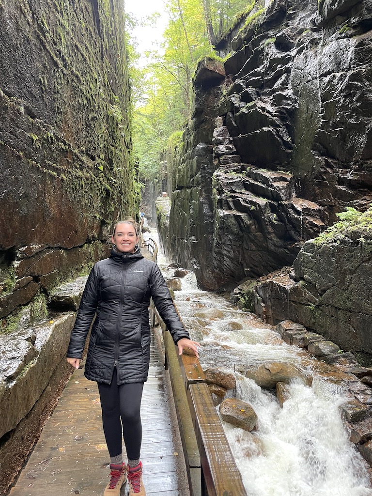 Woman in winter jacket on boardwalk in narrow, granite gorge next to flowing river and surrounded by rocky cliffs with lush vegetation. Flume Gorge, Franconia Notch State Park, White Mountains, New Hampshire.