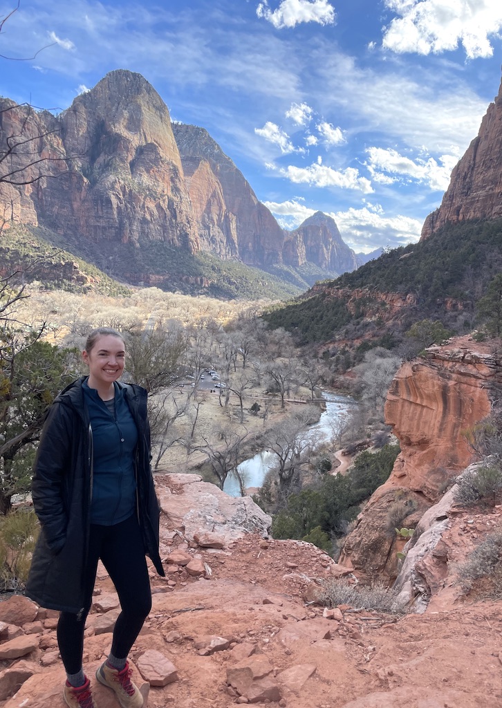Woman smiling at camera with dramatic views in background of huge red rock cliffs standing over valley with river winding through on sunny winter day. Hiking Middle Emerald Pools, Zion National Park, Utah.