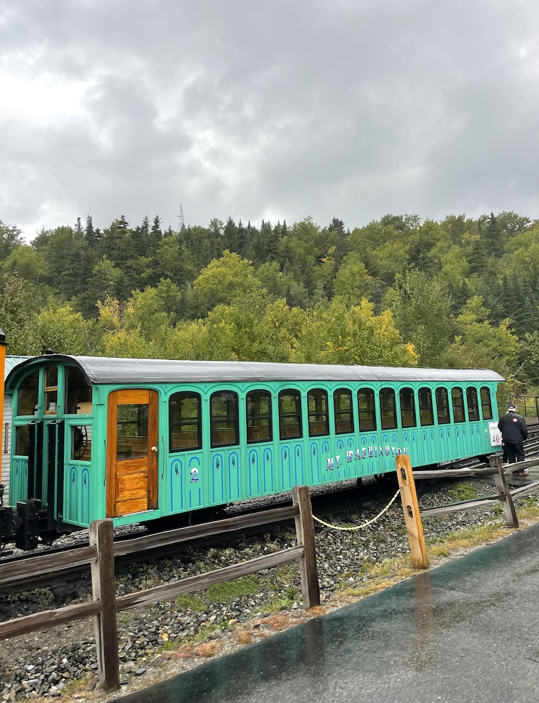 Green/ teal painted historic cog railway car on track with lush green trees beginning to turn yellow in background on cloudy, rainy early fall day. Mount Washington Cog Railway, White Mountains, New Hampshire.