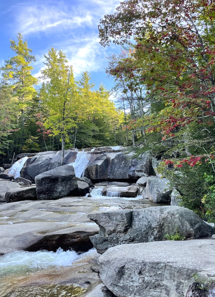 Small waterfalls and river flowing through very rocky river bed surrounded by lush green trees beginning to turn red and yellow in early fall on a sunny evening. Diana's Baths trail, White Mountains, New Hampshire.
