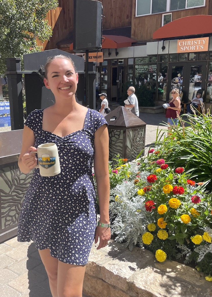 Woman in dress smiling and holding beer stein with flowers and shops in background on sunny early fall day. Vail Oktoberfest, Vail, Colorado.