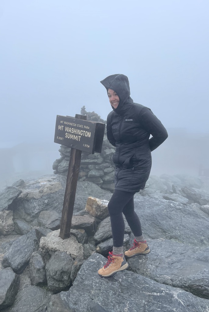 Woman standing on top summit of Mount Washington, she's on rocky surface next to small wooden sign reading "Mt Washington Summit", woman is in winter jacket with hood up, hood is blowing in the wind and there is almost no visibility due to thick clouds and fog. Mount Washington summit in early fall, White Mountains, New Hampshire.