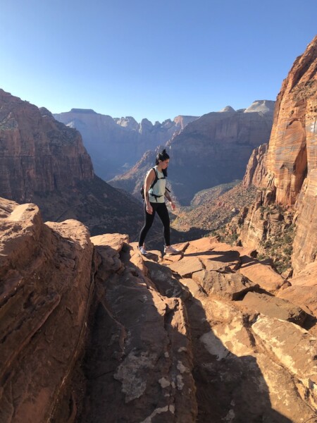 Woman hiking on red rock with dramatic red rock canyon views in background on sunny fall day. Canyon Overlook Trail, Zion Canyon, Zion National Park, Utah.