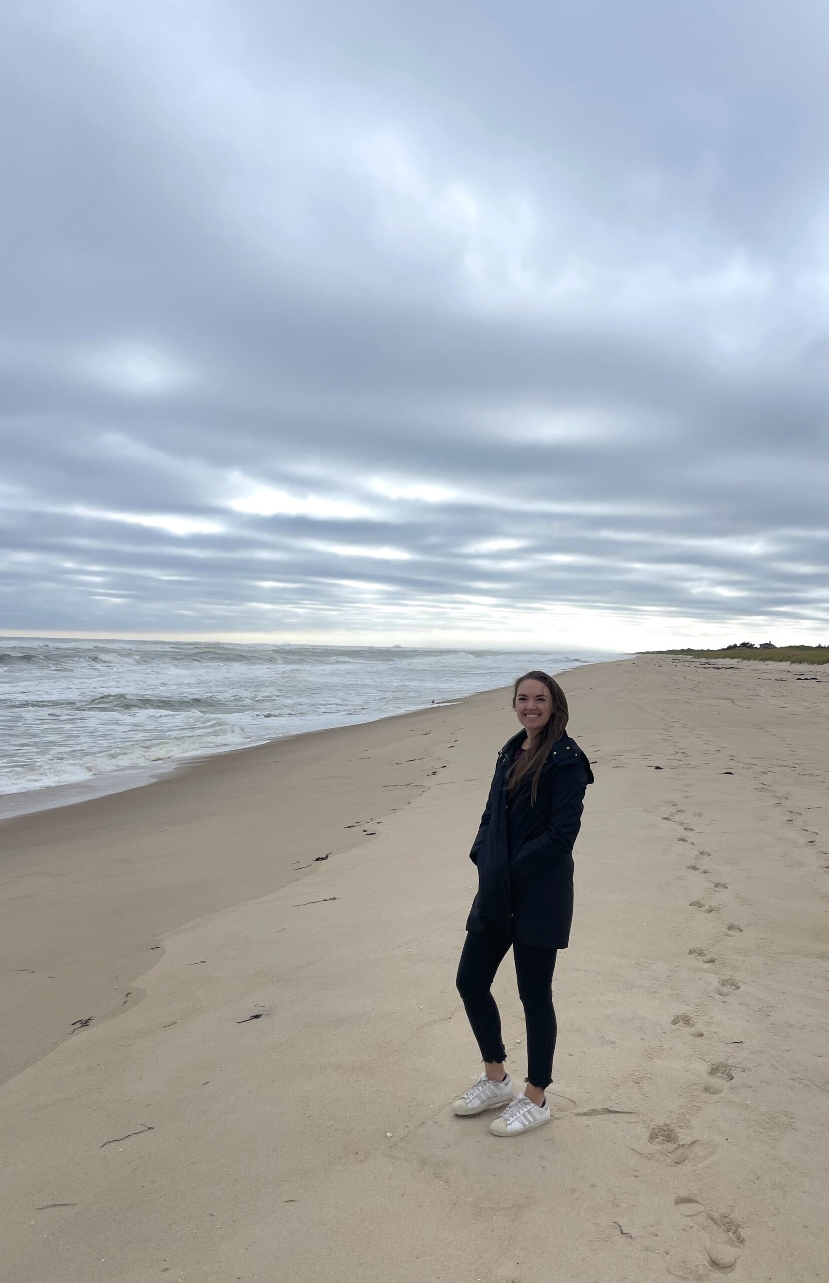 Woman in jacket and pants on golden sandy beach next to rough Atlantic Ocean on cloudy fall day. Atlantic Avenue Beach, Amagansett, New York, The Hamptons.